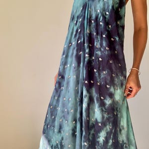 May include: A flowing, sleeveless dress in a tie-dye pattern of teal and navy blue, adorned with small, silver-colored embellishments. The dress features a halter neckline and a loose, comfortable fit, suitable for warm weather.