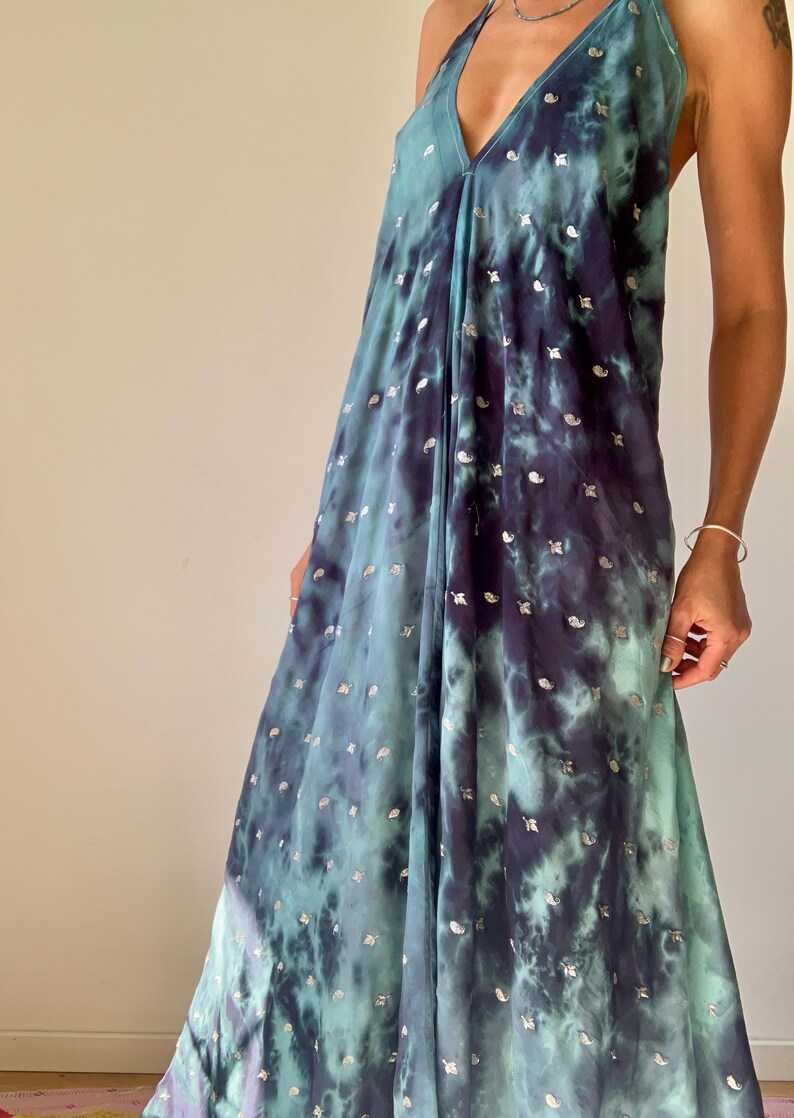 May include: A flowing, sleeveless dress in a tie-dye pattern of teal and navy blue, adorned with small, silver-colored embellishments. The dress features a halter neckline and a loose, comfortable fit, suitable for warm weather.