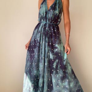 May include: A flowing, halter-neck maxi dress in shades of teal and deep purple, with a tie-dye effect. The dress features a cinched waist and is adorned with small, silver-colored embellishments. The dress is made of a lightweight fabric.