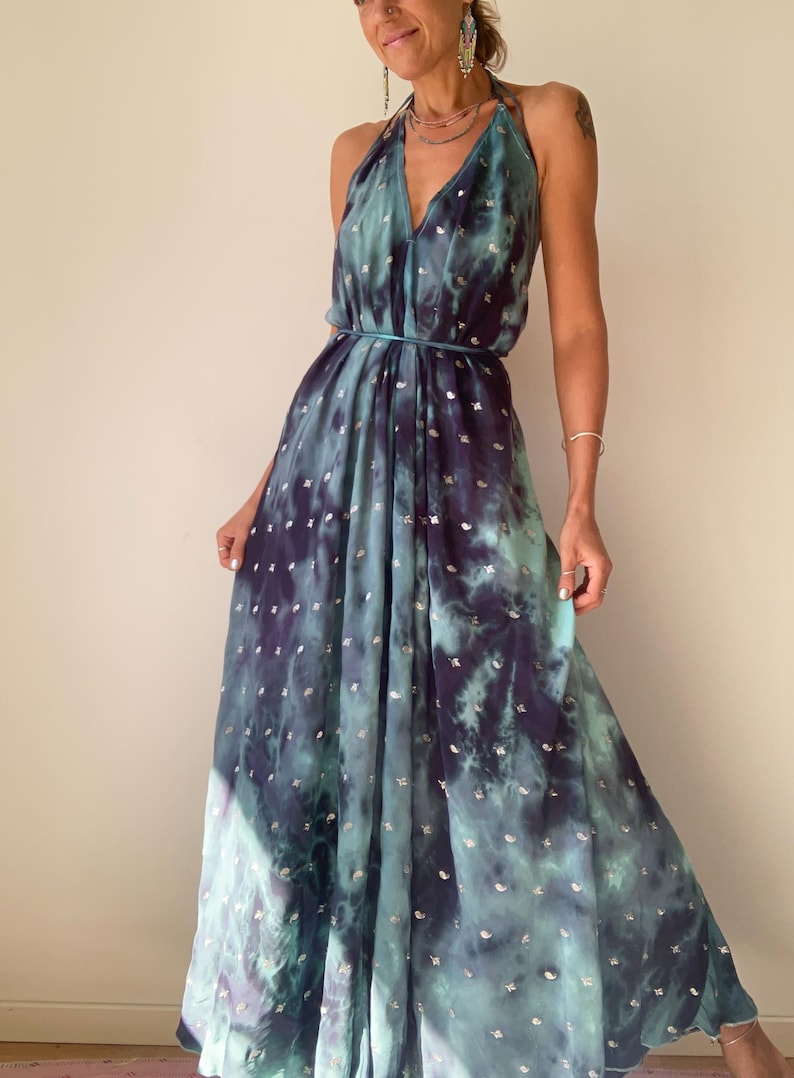 May include: A flowing, halter-neck maxi dress in shades of teal and deep purple, with a tie-dye effect. The dress features a cinched waist and is adorned with small, silver-colored embellishments. The dress is made of a lightweight fabric.
