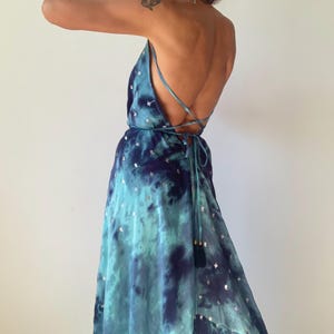May include: A floor-length dress in shades of teal and navy blue with a tie-dye pattern and small silver metallic accents. The garment features a backless design with thin spaghetti straps that cross and tie at the lower back.