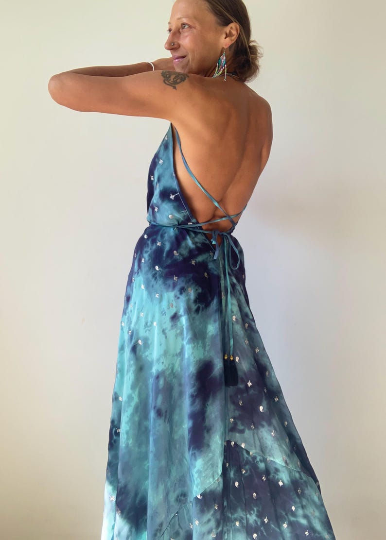 May include: A floor-length dress in shades of teal and navy blue with a tie-dye pattern and small silver metallic accents. The garment features a backless design with thin spaghetti straps that cross and tie at the lower back.