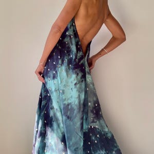 May include: A flowing, backless maxi dress in shades of teal, blue, and purple, with a tie-neck halter design. The dress features a tie-dye pattern and is embellished with small, silver-colored sequins scattered across the fabric. The dress is made of a lightweight material.