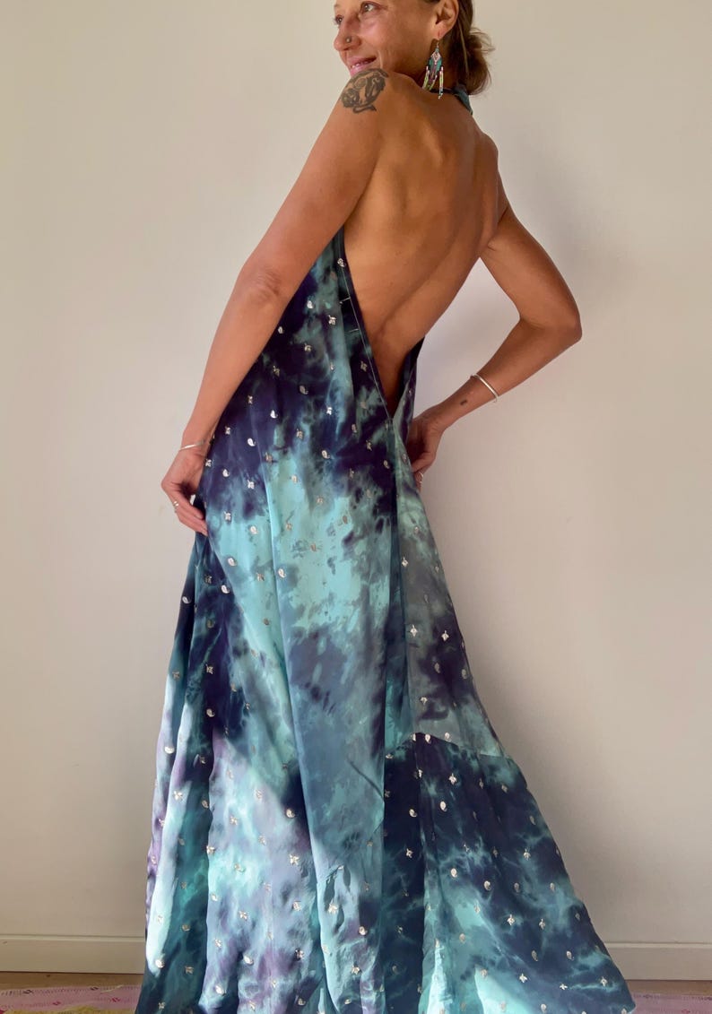 May include: A flowing, backless maxi dress in shades of teal, blue, and purple, with a tie-neck halter design. The dress features a tie-dye pattern and is embellished with small, silver-colored sequins scattered across the fabric. The dress is made of a lightweight material.
