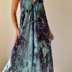 May include: A flowing, halter-neck maxi dress in shades of teal and deep indigo, with a tie-dye effect. The dress is adorned with small, silver-colored embellishments scattered across the fabric. The dress has a V-neckline and a loose, comfortable fit.