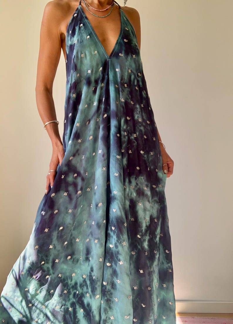 May include: A flowing, halter-neck maxi dress in shades of teal and deep indigo, with a tie-dye effect. The dress is adorned with small, silver-colored embellishments scattered across the fabric. The dress has a V-neckline and a loose, comfortable fit.