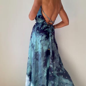 May include: A flowing, tie-dye maxi dress in shades of blue and teal, adorned with silver star-shaped embellishments. The dress features a low back and spaghetti straps that crisscross. The dress is floor-length and has a relaxed fit.