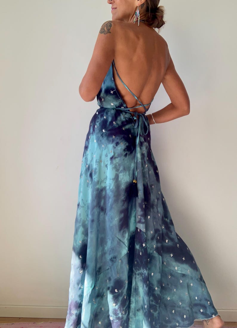 May include: A flowing, tie-dye maxi dress in shades of blue and teal, adorned with silver star-shaped embellishments. The dress features a low back and spaghetti straps that crisscross. The dress is floor-length and has a relaxed fit.