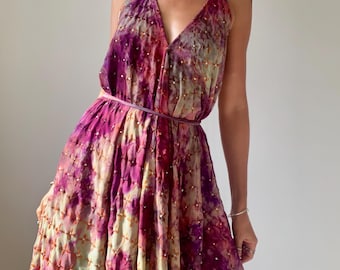 May include: A sleeveless halter-neck maxi dress featuring a tie-dye pattern in shades of deep purple, magenta, and pale yellow. The fabric is adorned with a grid of small, embroidered cross-stitch details and a thin, matching fabric waist tie.