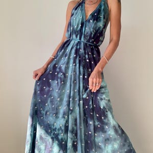 May include: A floor-length halter dress featuring a deep blue and teal tie-dye pattern with small silver metallic accents throughout. The garment has a plunging V-neckline, a thin waist tie, and a flowing, pleated skirt that reaches the floor.