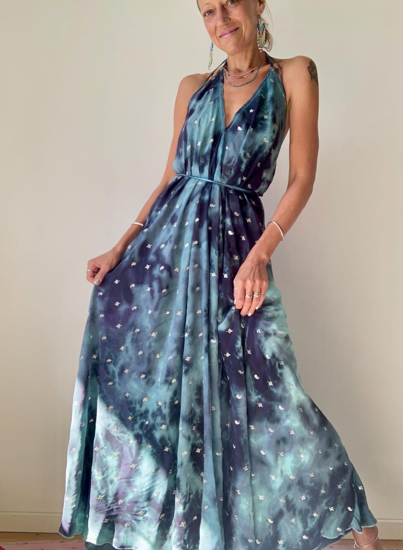 May include: A floor-length halter dress featuring a deep blue and teal tie-dye pattern with small silver metallic accents throughout. The garment has a plunging V-neckline, a thin waist tie, and a flowing, pleated skirt that reaches the floor.
