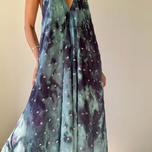 May include: A flowing, halter-neck maxi dress in shades of teal and deep purple, with a tie-dye effect. The dress is adorned with small, silver-colored embellishments scattered across the fabric. The dress has a V-neckline and a loose, comfortable fit.
