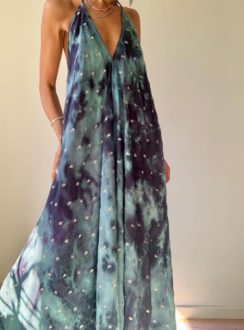May include: A flowing, halter-neck maxi dress in shades of teal and deep purple, with a tie-dye effect. The dress is adorned with small, silver-colored embellishments scattered across the fabric. The dress has a V-neckline and a loose, comfortable fit.