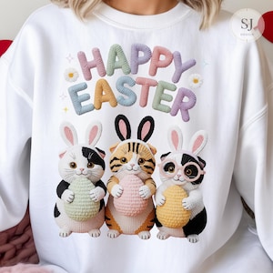 May include: White sweatshirt with a colourful "HAPPY EASTER" design. The graphic features three cartoon cats wearing bunny ears and holding Easter eggs. The cats are black and white, orange and brown, and black and white with glasses.