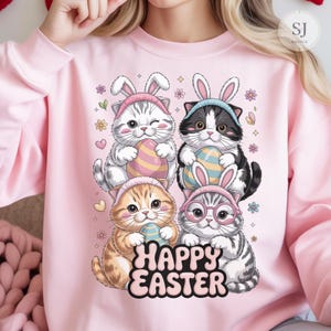 May include: A pink sweatshirt with a design of four cartoon kittens wearing bunny ears and holding Easter eggs. The words "HAPPY EASTER" are printed below the kittens. The design includes floral and heart accents.