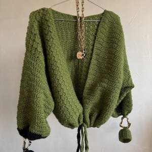 May include: A handmade olive green crochet cardigan with a V-neck design. The cardigan features black cuffs and a drawstring closure. A gold-tone necklace with a pendant hangs from the hanger. The cardigan has a unique, textured stitch pattern.