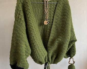 Puede incluir: Cárdigan de crochet verde oliva hecho a mano con diseño de cuello en V. El cárdigan presenta puños negros y un cierre de cordón. Un collar dorado con un colgante cuelga de la percha. El cárdigan tiene un patrón de puntada único y texturizado.