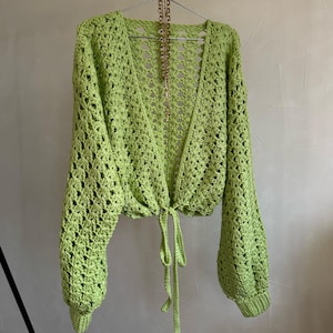 May include: A lime green, open-weave crochet cardigan with long sleeves and a tie closure at the waist. The cardigan is hanging on a metal hanger. A gold-colored necklace is draped over the hanger.
