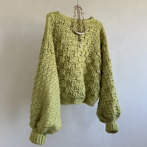 May include: A lime green long-sleeve sweater featuring a chunky crochet basketweave texture, a round neckline, and ribbed cuffs. The garment is displayed on a metal hanger with a gold-toned chain necklace draped over the front neckline area.