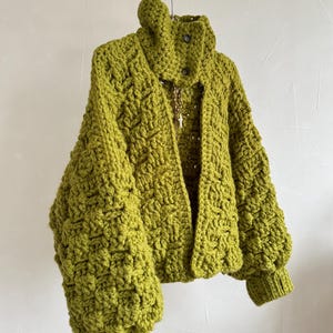 Puede incluir: Chaqueta de ganchillo verde oliva, con cuello alto y cierre de botones. La chaqueta presenta un patrón de puntadas texturizadas y mangas voluminosas. Un collar dorado con un colgante de cruz cuelga dentro de la parte delantera abierta.
