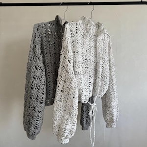 May include: Two long-sleeved crochet sweaters hanging on hangers. One sweater is solid gray, and the other is white with small black speckles. Both garments feature a loose, open-knit floral pattern, high necklines, and tie-front closures.