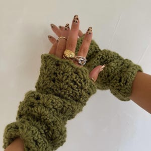 Puede incluir: Guantes sin dedos de ganchillo verde oliva. Los guantes están hechos de un hilo grueso y tienen un patrón texturizado. Los guantes se muestran con múltiples anillos en los dedos.