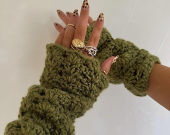 May include: Olive green crochet fingerless gloves. The gloves are made of a chunky yarn and have a textured pattern. The gloves are shown with multiple rings on the fingers.