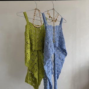 May include: Two open-weave crochet tops in lime green and light blue hang on a metal rod. Gold necklaces are draped over the tops. The tops have a sleeveless design with a flowing, asymmetrical hemline.