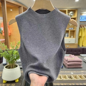May include: A grey sleeveless knitted top with a high neck. The top is made of ribbed knit fabric and is displayed on a wooden hanger. The background includes a plant in a white pot and other clothing items.