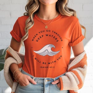 May include: An orange short-sleeved t-shirt featuring a graphic of a stylized wave in the center. The text reads "WHEN YOU GO THROUGH DEEP WATERS I WILL BE WITH YOU" in a circular arrangement, with "ISAIAH 43:2" printed in black below the wave.