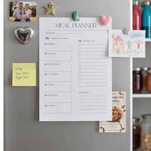 Pode incluir: Um planejador de refeições branco com o texto "MEAL PLANNER" e seções para refeições semanais e uma lista de tarefas. O planejador é preso a uma geladeira com ímãs, incluindo uma foto de família e um ímã em forma de coração.