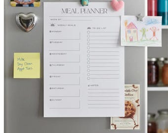 May include: A white meal planner with the text "MEAL PLANNER" and sections for weekly meals and a to-do list. The planner is held on a refrigerator with magnets, including a family photo and a heart-shaped magnet.