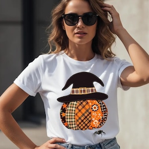 Pode incluir: Camiseta branca com um design de Halloween. O gráfico inclui um chapéu de bruxa preto sobre uma abóbora de retalhos laranja, verde e marrom. Uma pequena aranha preta está embaixo.