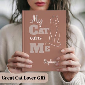 Könnte beinhalten: Ein terrakottafarbenes Notizbuch mit festem Einband, das den weißen Schriftzug "My Cat owns Me" und eine minimalistische Strichzeichnung einer sitzenden Katze zeigt. Der Name "Stephanie" ist in weißer Schreibschrift unten rechts aufgedruckt.
