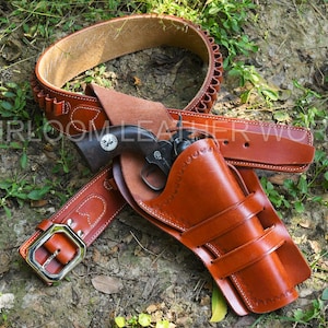 Könnte beinhalten: Ein braunes Leder-Pistolenholster mit passendem Gürtel und Patronenhalter. Das Holster ist für einen Revolver konzipiert, und der Gürtel hat eine große silberne Schnalle. Das Set wird auf grünem Laubwerk präsentiert.