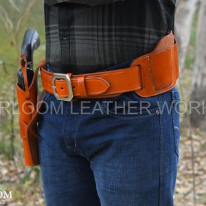Pode incluir: Um cinto de couro castanho com coldre a condizer, usado sobre calças de ganga azuis. O cinto tem uma fivela prateada grande e uma parte traseira larga. O coldre contém um revólver preto. O texto "HEIRLOOM LEATHER WORKS" é visível.