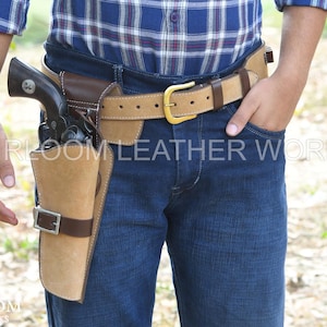 May include: A tan leather holster with a brown leather accent and a silver buckle, holding a black revolver. The holster is attached to a tan and brown leather belt with a gold buckle. The text "HEIRLOOM LEATHER WORKS" is visible.