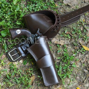Könnte beinhalten: Ein braunes Lederholster-, Gürtel- und Pistolen-Set. Das Holster ist für einen Revolver konzipiert. Der Gürtel hat eine silberne Schnalle. Die Pistole hat einen Holzgriff. Das Set wird auf einem Bett aus grünen Pflanzen und Erde präsentiert.