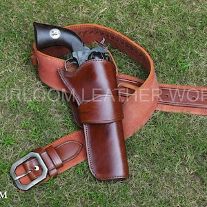 Könnte beinhalten: Ein braunes Lederholster- und Gürtelset mit einer silbernen Schnalle, das einen Revolver hält. Das Set wird auf grünem Gras präsentiert. Der Text "HEIRLOOM LEATHER WORKS" ist sichtbar.