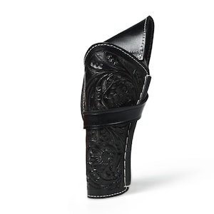 May include: A black leather holster with intricate floral and scroll tooling. The holster features white stitching along the edges and a strap. The design is a classic Western style, suitable for carrying a firearm.