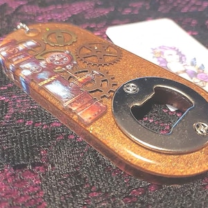 Puede incluir: Un abridor de botellas con temática steampunk con acabado metálico. El abridor tiene un cuerpo marrón de forma ovalada con engranajes incrustados y una ilustración detallada. Se adjunta un abridor de botellas plateado. Una pequeña etiqueta con una ilustración floral está en el fondo.