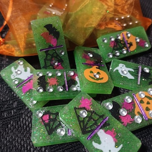 May include: Green glitter dominoes with Halloween designs. Each domino features a different spooky image, including bats, ghosts, spiders, and jack-o'-lanterns. The dominoes have silver dot markers and are displayed near an orange organza bag.