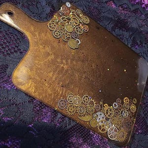 Puede incluir: Tabla de servir con temática steampunk y asa. La tabla es de color marrón oscuro con purpurina dorada y está decorada con varios engranajes, llaves y piezas de reloj de latón. El asa tiene un agujero para colgar.