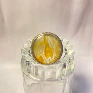 Könnte beinhalten: Ein runder, dekorativer Ring mit silberfarbenem Rand und einem gelb-weißen Marmormuster. Der Ring liegt auf einem transparenten, strukturierten Glasbehälter.