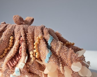 May include: A peach-colored, embellished clutch with a bow closure. The clutch is adorned with sequins, beads, and light blue accents. The bottom edge features a row of large, round, pale peach-colored sequins.