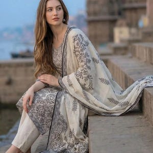 Può includere: Un completo color crema in due pezzi con una sciarpa abbinata, con un motivo floreale grigio scuro. Il completo comprende un top a maniche lunghe e pantaloni. La sciarpa è drappeggiata sulle spalle. Il completo è indossato con sandali.