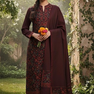 May include: A burgundy salwar kameez ensemble featuring a floral print. The outfit includes a long-sleeved tunic, matching pants, and a shawl. The tunic has detailed embroidery around the neckline and cuffs. The person is holding a small bouquet of flowers.