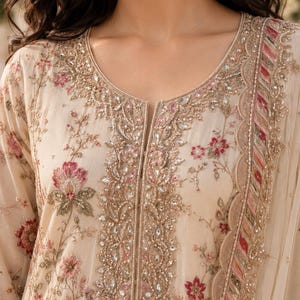 May include: A beige tunic featuring intricate floral embroidery in shades of pink and green, accented with gold threadwork and clear crystals along the neckline and front placket, paired with a matching dupatta with a scalloped edge.