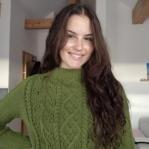 Könnte beinhalten: Ein grüner Langarmpullover mit hohem Ausschnitt. Der Pullover hat ein detailliertes Zopfmuster auf der Vorderseite und an den Ärmeln. Die Farbe ist ein leuchtendes, erdiges Grün, und der Pullover scheint aus einem weichen, warmen Material zu sein.
