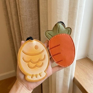 Puede incluir: Dos llaveros de cuero: uno con forma de pez en tonos amarillos y naranjas, y otro de zanahoria en naranja y verde. Ambos tienen lazos negros para sujetar las llaves. El llavero de pez tiene un diseño detallado.